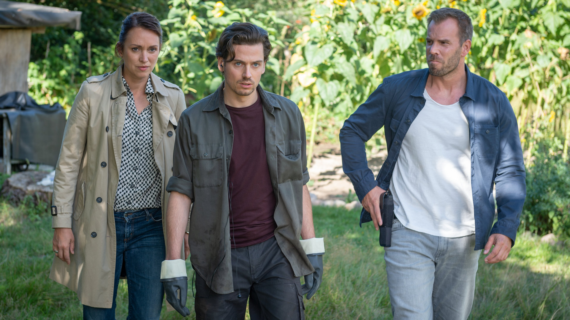 "SOKO Wismar – Totenruhe": Auf dem Gelände des Bestattungsinstituts geht Karoline Joost (Nike Fuhrmann) neben ihrem Kollegen Lars Pöhlmann (Dominic Boeer). Vor ihnen geht Nils Berger (Anthony Paul). Beide Kommissare haben ihn fest im Blick. Lars Pöhlmann hat seine Hand an seiner Waffe.