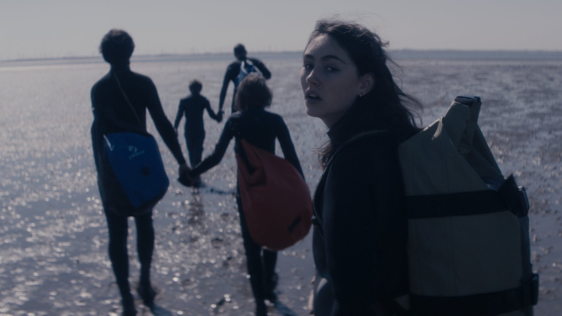 "Sløborn - Weg hier": Evelin (Emily Kusche) und ihre Freunde laufen übers Watt. Emily trägt einen Rucksack und sieht erschöpft in die Kamera.