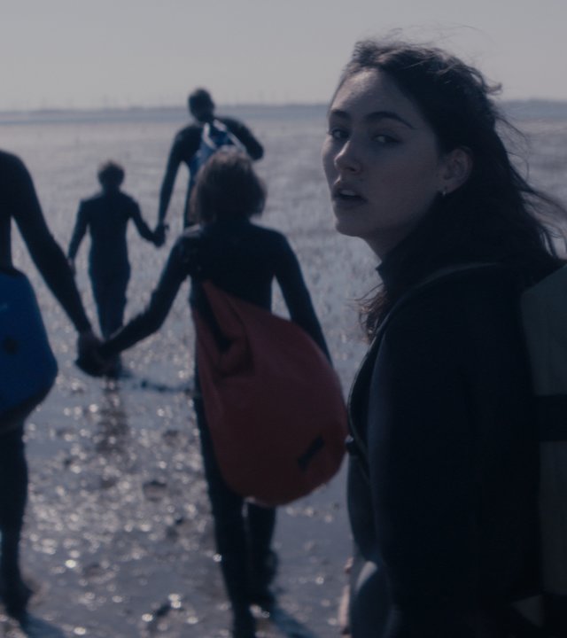 "Sløborn - Weg hier": Evelin (Emily Kusche) und ihre Freunde laufen übers Watt. Emily trägt einen Rucksack und sieht erschöpft in die Kamera.