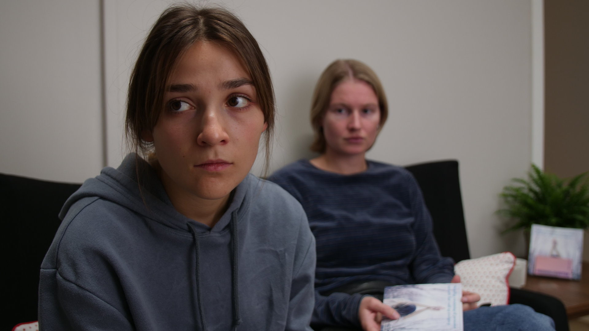 "About Saturday - Hätte Nein sagen können": Lotte (Hanna Heider Hov) sitzt hinter Klara (Darin Hagi) auf einem Sofa und schaut sie an, während Klara geistesabwesend in die Ferne blickt.