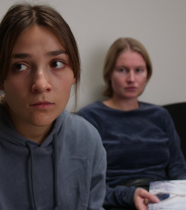 "About Saturday - Hätte Nein sagen können": Lotte (Hanna Heider Hov) sitzt hinter Klara (Darin Hagi) auf einem Sofa und schaut sie an, während Klara geistesabwesend in die Ferne blickt.