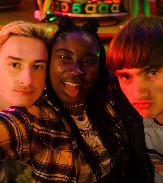 "Tell me everything: Alles ist okay": Jonny (Eden H. Davies), Neve (Lauryn Ajufo) und Louis (Spike E. Fearn) stehen vor dem Fahrgeschäft einer Kirmes und blicken in die Kamera, um ein Selfie zu machen.