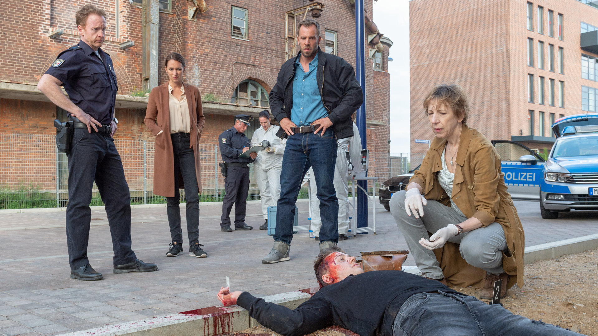 „SOKO Wismar – Zu Tode erschreckt“: Jannik Claasen (Felix Jordan) liegt rücklings und blutüberströmt an einem sandigen Bürgersteig. Bei ihm kniet Helene Sturbeck (Katharina Blaschke). Die Kollegen Timmermann (Mathias Junge), Joost (Nike Fuhrmann) und Pöhlmann (Dominic Boeer) stehen am Kopfende des Toten und blicken zu ihm. Im Hintergrund parkt ein Polizeiauto vor Backsteinhäusern. Kollegen in Schutzanzügen verrichten ihre Arbeit.