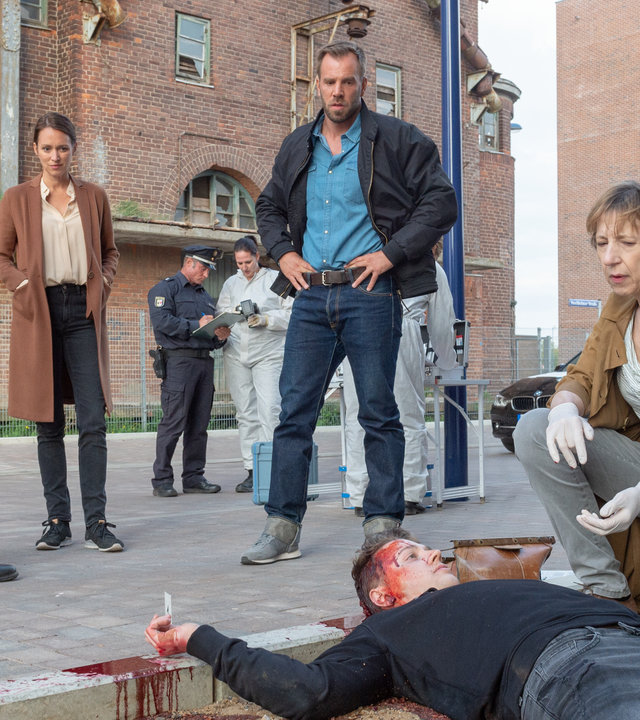 „SOKO Wismar – Zu Tode erschreckt“: Jannik Claasen (Felix Jordan) liegt rücklings und blutüberströmt an einem sandigen Bürgersteig. Bei ihm kniet Helene Sturbeck (Katharina Blaschke). Die Kollegen Timmermann (Mathias Junge), Joost (Nike Fuhrmann) und Pöhlmann (Dominic Boeer) stehen am Kopfende des Toten und blicken zu ihm. Im Hintergrund parkt ein Polizeiauto vor Backsteinhäusern. Kollegen in Schutzanzügen verrichten ihre Arbeit.