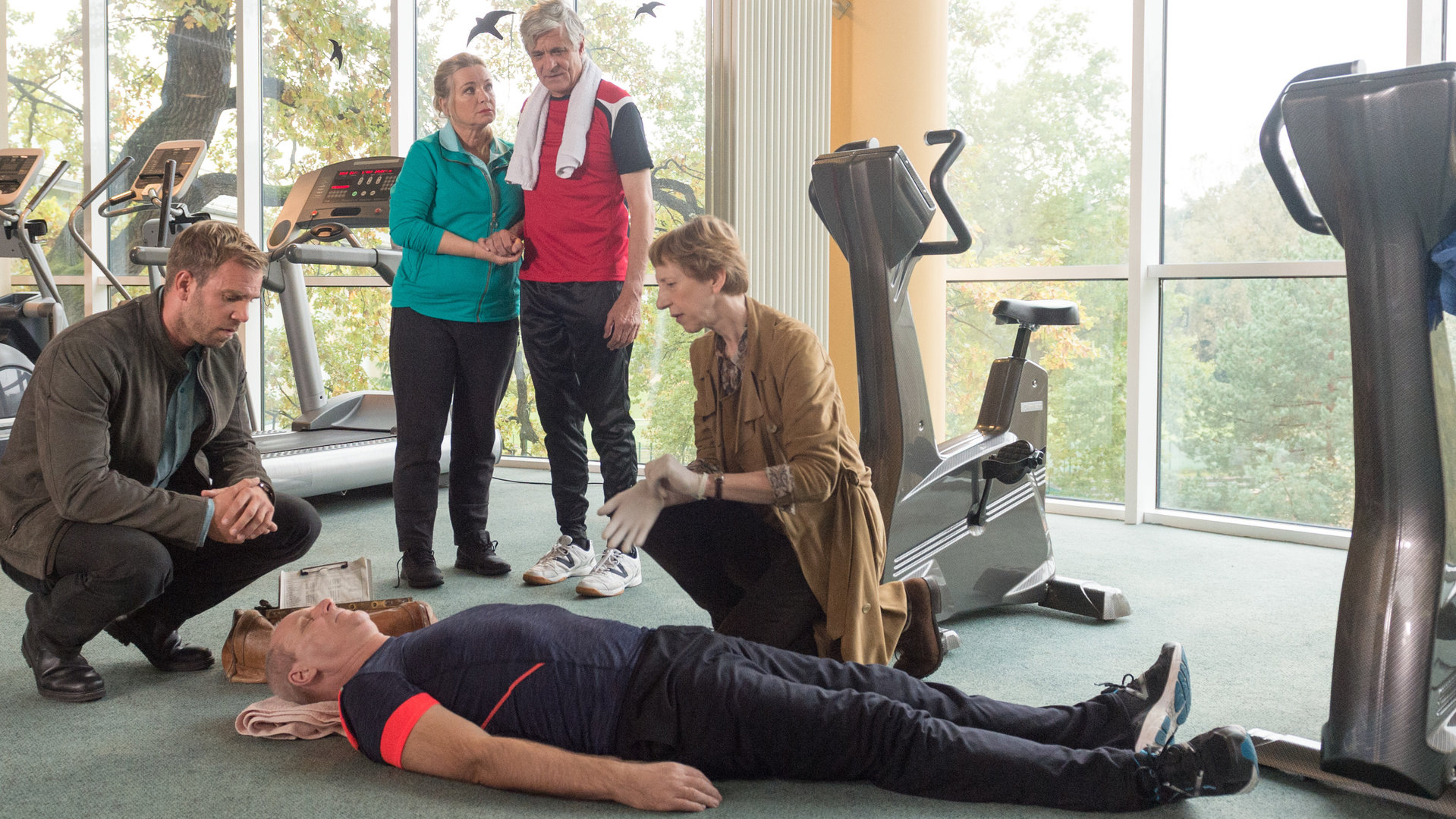 „SOKO Wismar – Fit für die Ewigkeit“: Im Fitnessraum liegt Markus Rotmanns (Herbert Trattnigg) lebloser Körper  am Boden. Um ihn herum sind Rechtsmedizinerin Helene Sturbeck (Katharina Blaschke) und Kommissar Lars Pöhlmann (Dominic Boeer), im Hintergrund stehen Hans (Dietrich Adam) und Elisabeth Güstrow (Susanne Bentzien).