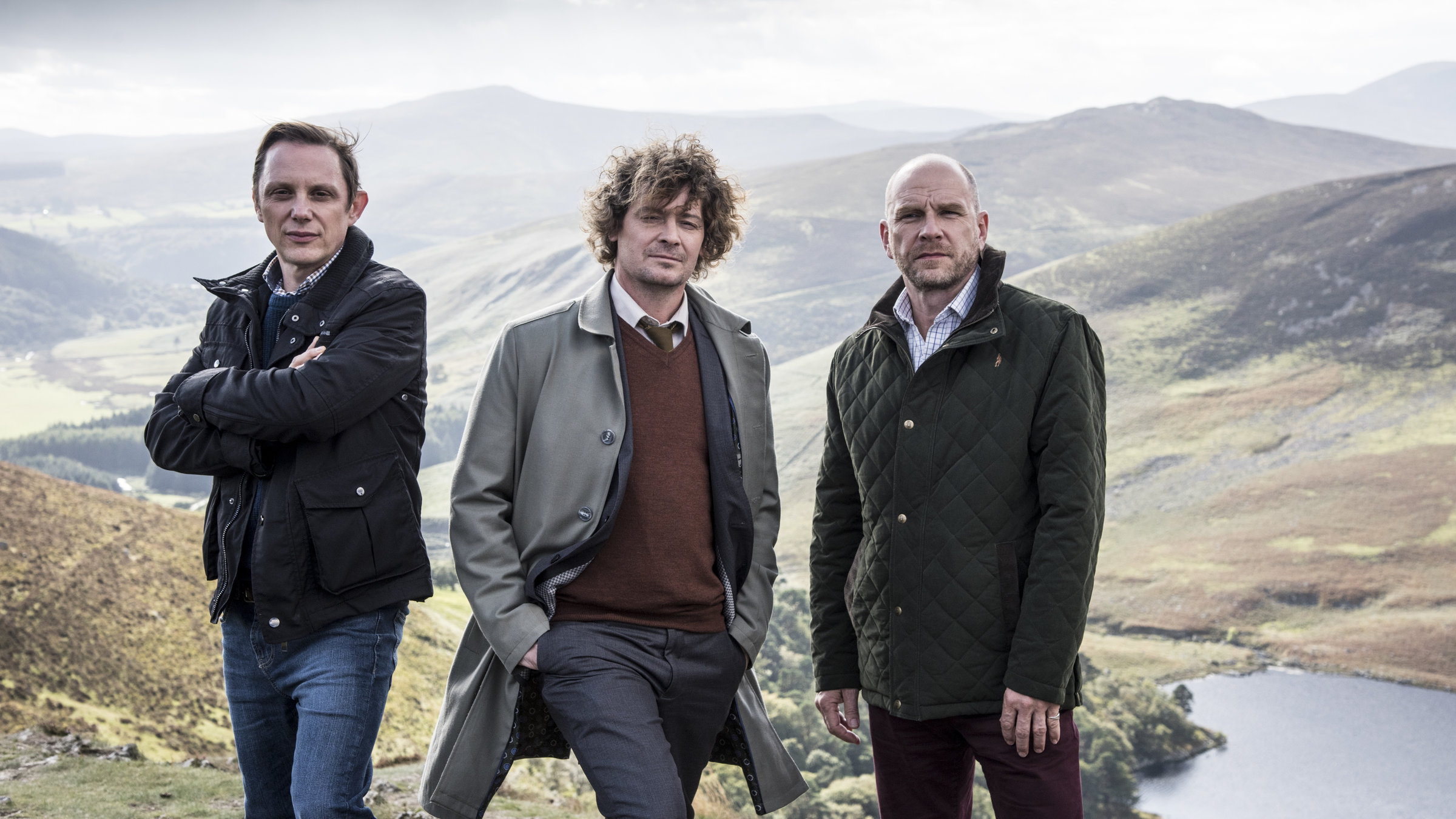 "Inspektor Jury - Mord im Nebel":  Assistent Alfred Wiggins (Arndt Schwering-Sohnrey), Inspektor Jury (Fritz Karl) mit beiden Händen in der Hosentasche und der adlig dreinschauend Melrose Plant (Götz Schubert), im Hintergrund ein weites Bergtal unter dramatisch bewölktem Himmel.