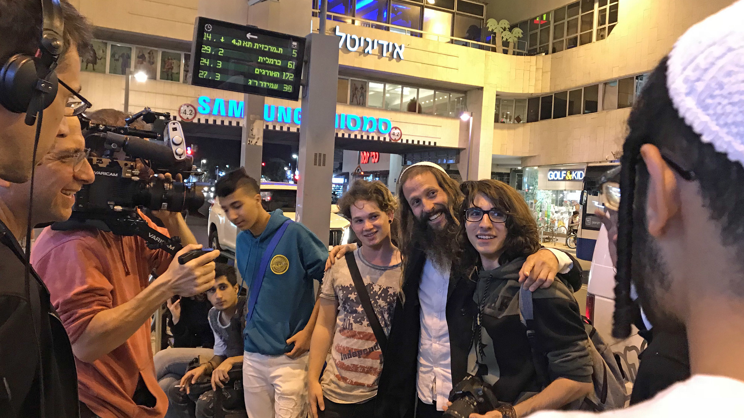 "Tel Aviv - Lebensfreude trotz allem": Ein orthodoxer Hippie steht zwischen zwei Jugendlichen in einem Gebäude und hat seine Arme um deren Schultern gelegt. Die drei lachen einem Mann zu, der nur von hinten zu sehen ist. Ein Kamerateam filmt das Geschehen.