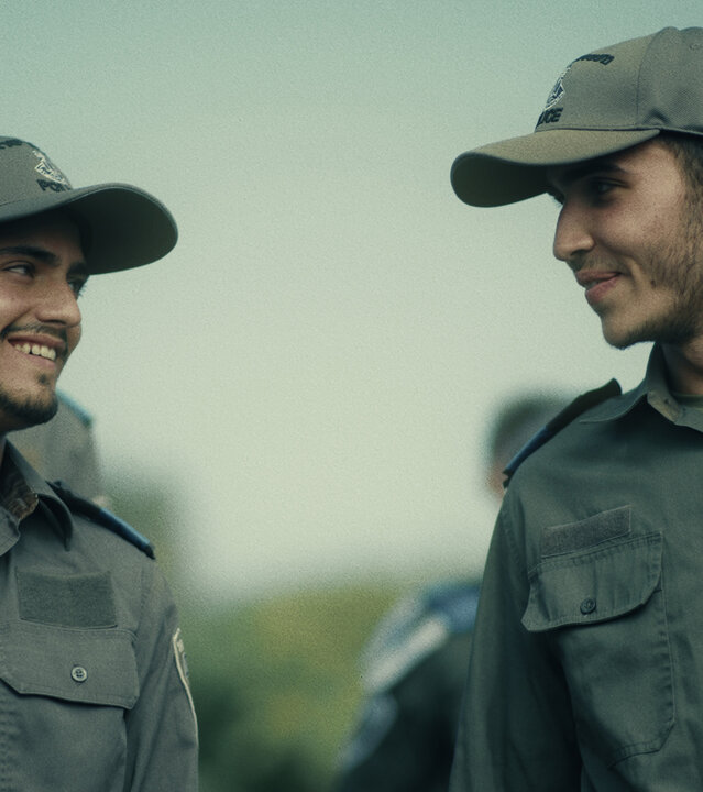 "Borders - Wut": Kobi (Ido Elieli) und Avi (Ben Sultan) stehen nebeneinander. Sie tragen ihre Uniform und Kappen. Beide schauen sich gegenseitig an und lächeln.