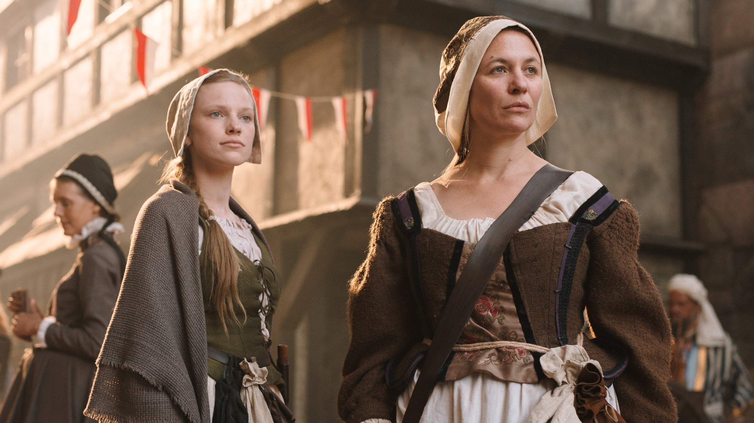 "Terra X: Ein Tag in Köln 1629": Spielszene: Hebamme Anna Stein (Julia Thurnau) steht neben ihrer Lehrmagd (Elina Vaska) auf der Straße. Beide tragen mittelalterliche Gewänder und eine Haube auf dem Kopf. An ihrem Gürtel hängt jeweils ein Beutel.