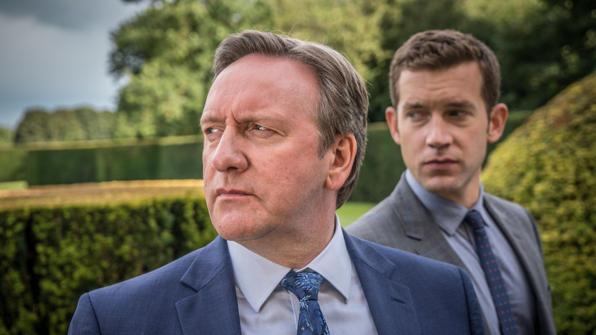 "Inspector Barnaby - Eine Hochzeit und dreieinhalb Todesfälle": DCI John Barnaby (Neil Dudgeon) und sein Assistent DS Jamie Winter (Nick Hendrix) stehen zwischen sauber geschnittenen Hecken in einem parkähnlichen Garten und schauen nach links.