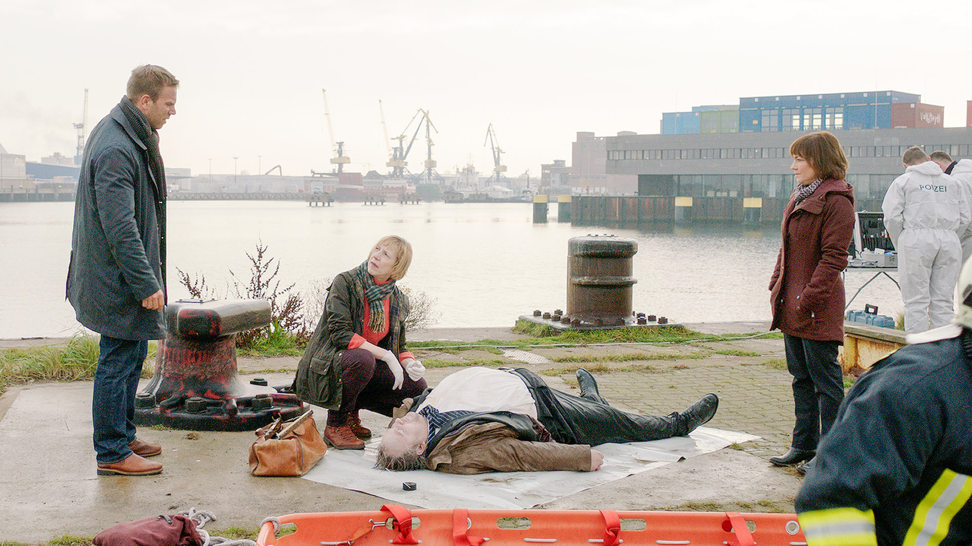 "SOKO Wismar – Der Dicke": Lars Pöhlmann (Dominic Boeer) und Katrin Börensen (Claudia Schmutzler) stehen am Hafen bei der Pathologin Helene Sturbeck (Katharina Blaschke), die die durchnässte Leiche eines kräftigen Mannes untersucht. Im Hintergrund sind die Hafenkräne zu sehen.