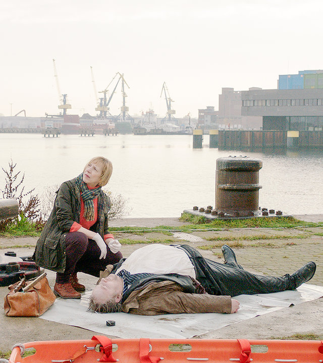 "SOKO Wismar – Der Dicke": Lars Pöhlmann (Dominic Boeer) und Katrin Börensen (Claudia Schmutzler) stehen am Hafen bei der Pathologin Helene Sturbeck (Katharina Blaschke), die die durchnässte Leiche eines kräftigen Mannes untersucht. Im Hintergrund sind die Hafenkräne zu sehen.