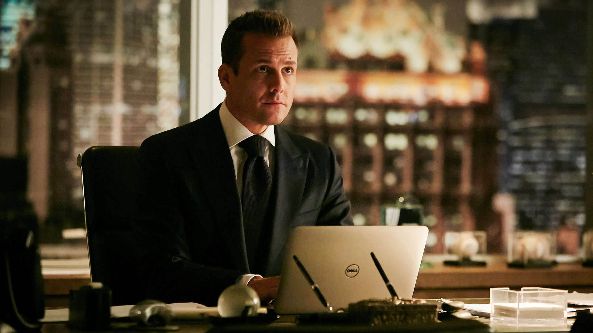 "Suits - Das Versprechen": Harvey (Gabriel Macht)  sitzt am Schreibtisch vor seinem aufgeklappten Laptop und schaut interessiert. Im Hintergrund sind die bei Abend erleuchteten Hochhäuser New Yorks zu erkennen.