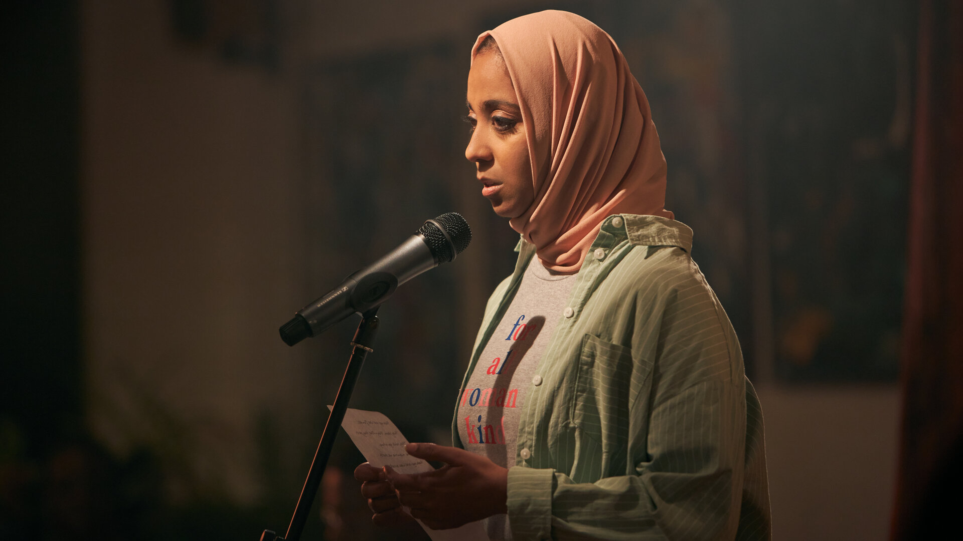 "We are Lady Parts - Godzilla": Amina (Anjana Vasan) steht vor einem Mikrofon und hält ein Blatt Papier in der Hand. Sie trägt ein rosafarbenes Kopftuch, ein gestreiftes Hemd über einem T-Shirt und blickt konzentriert geradeaus.