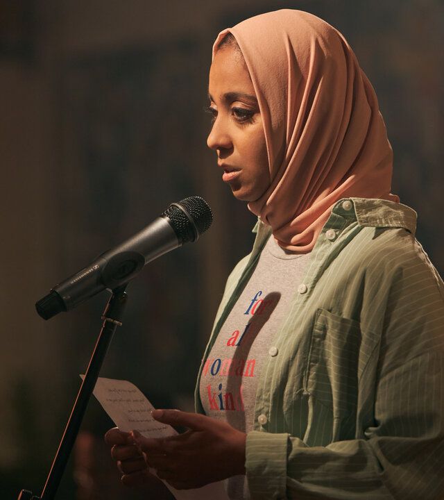 "We are Lady Parts - Godzilla": Amina (Anjana Vasan) steht vor einem Mikrofon und hält ein Blatt Papier in der Hand. Sie trägt ein rosafarbenes Kopftuch, ein gestreiftes Hemd über einem T-Shirt und blickt konzentriert geradeaus.