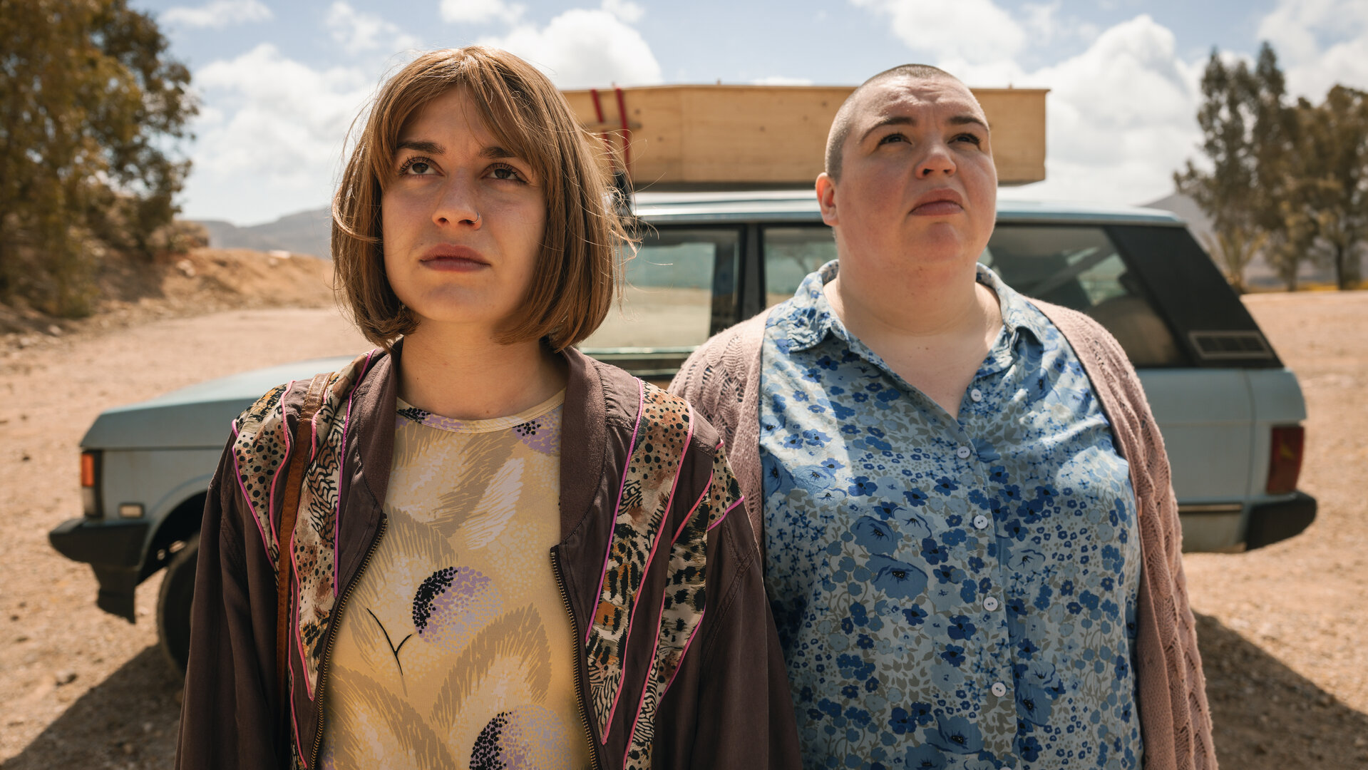 "How to Kill Your Sister - Folge 4": Anna (Emma Rotsaert) und Kat (Marjan De Schutter) stehen im Freien nebeneinander. Im Hintergrund parkt ein altes Auto mit einem Holzsarg auf dem Dach. Anna trägt eine Perücke mit mittellangen dunkelblonden Haaren. Kat trägt ihre Haare raspelkurz.