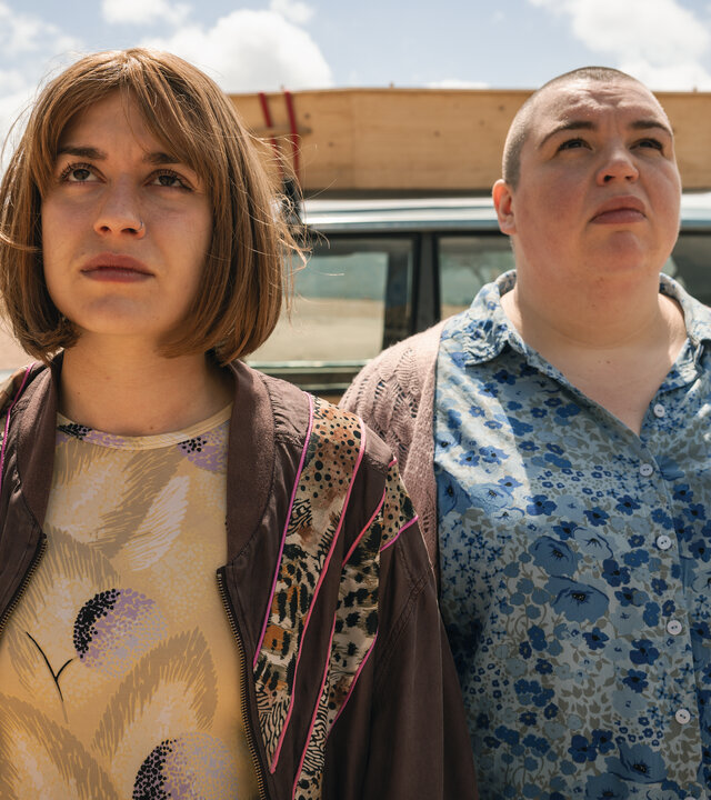 "How to Kill Your Sister - Folge 4": Anna (Emma Rotsaert) und Kat (Marjan De Schutter) stehen im Freien nebeneinander. Im Hintergrund parkt ein altes Auto mit einem Holzsarg auf dem Dach. Anna trägt eine Perücke mit mittellangen dunkelblonden Haaren. Kat trägt ihre Haare raspelkurz.