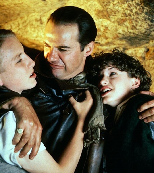 "Fünf Freunde machen eine Entdeckung": Der in einer schwarzen Lederjacke gekleidete Emilio (Paul Herzberg) hat seinen rechten Arm um Dicks (Paul Child) Hals und Schulter gelegt, während er ihn grimmig anschaut. In seinem linken Arm hält er George (Jemima Rooper) fest, die ihn anschaut. Die drei stehen dabei in einer Steinhöhle.