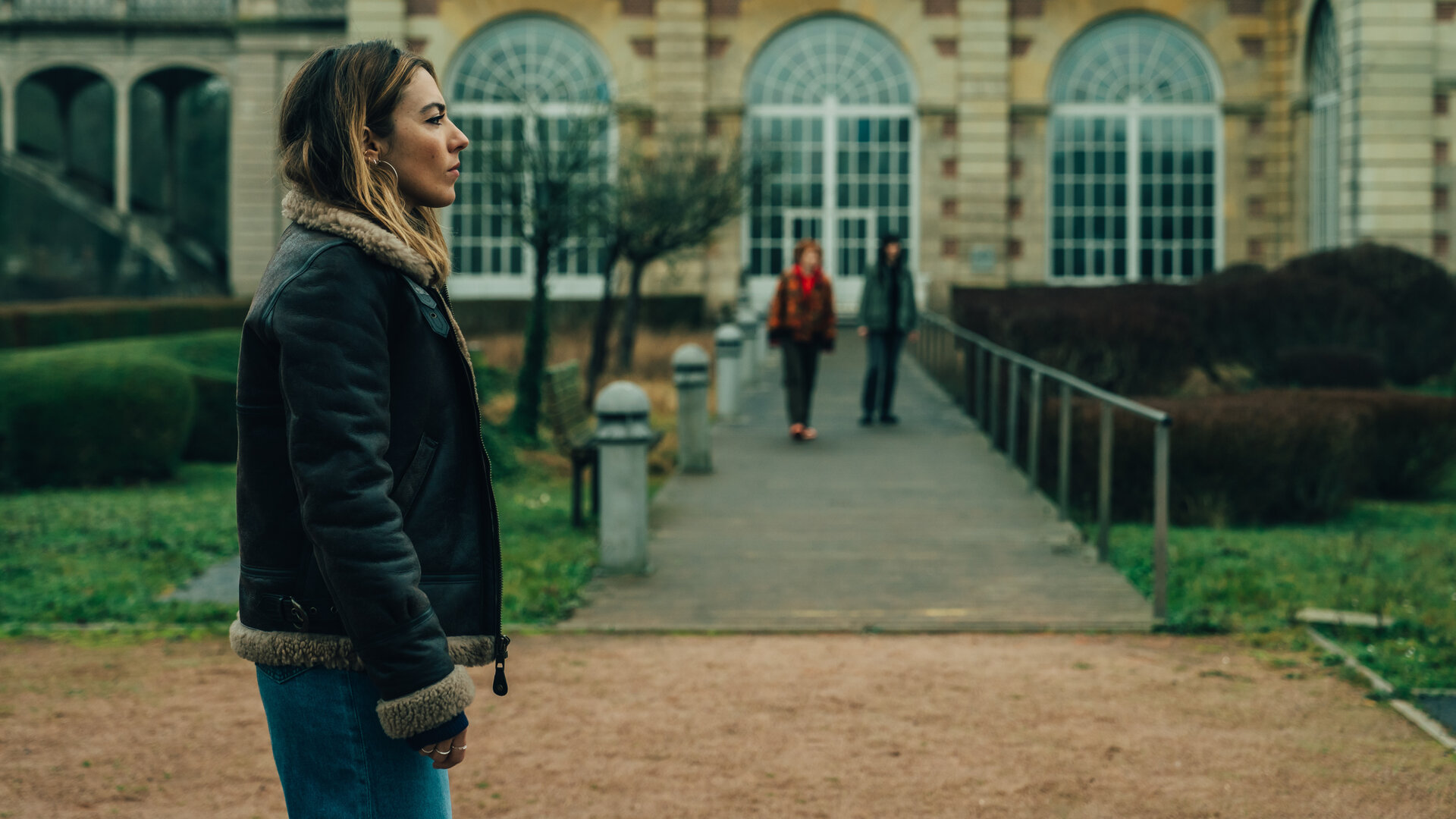 "Cat's Eyes - Sylia": Sylia (Constance Labbé) steht vor einem alten Gebäude mit großen Fenstern. Sie blickt zur Seite. Im Hintergrund sind unscharf Alexia (Claire Romain) und Tamara (Camille Lou) zu erkennen.