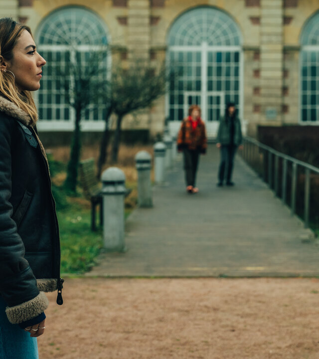 "Cat's Eyes - Sylia": Sylia (Constance Labbé) steht vor einem alten Gebäude mit großen Fenstern. Sie blickt zur Seite. Im Hintergrund sind unscharf Alexia (Claire Romain) und Tamara (Camille Lou) zu erkennen.