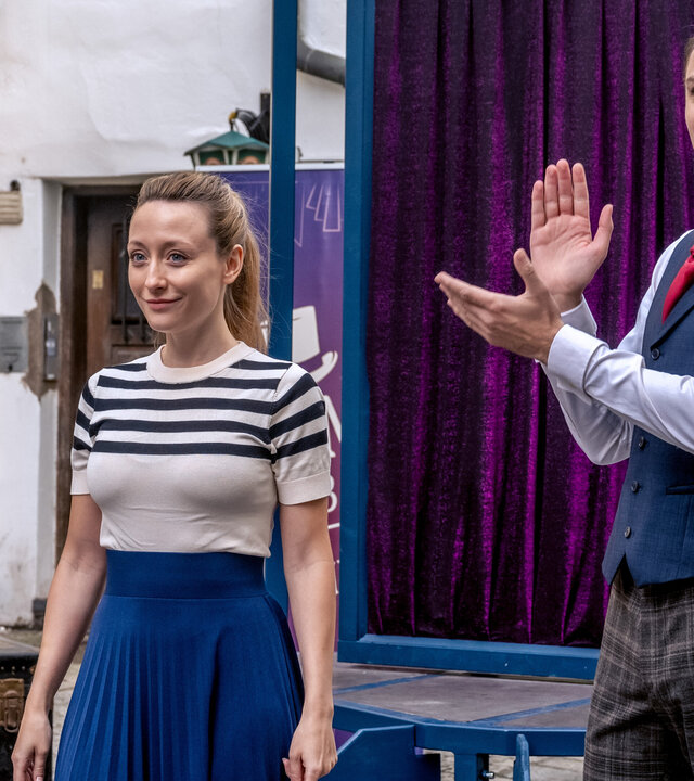 "SOKO Linz - Magic Moment": Anna Gruber (Alina Fritsch) und Sam Schubert (Michael Glantschnig) befinden sich in der Innenstadt. Hinter ihnen befindet sich eine mobile Bühne, auf der ein lila Samtvorhang angerbracht ist. Sam klatscht in die Hände, während Anna lächelnd neben ihm steht.