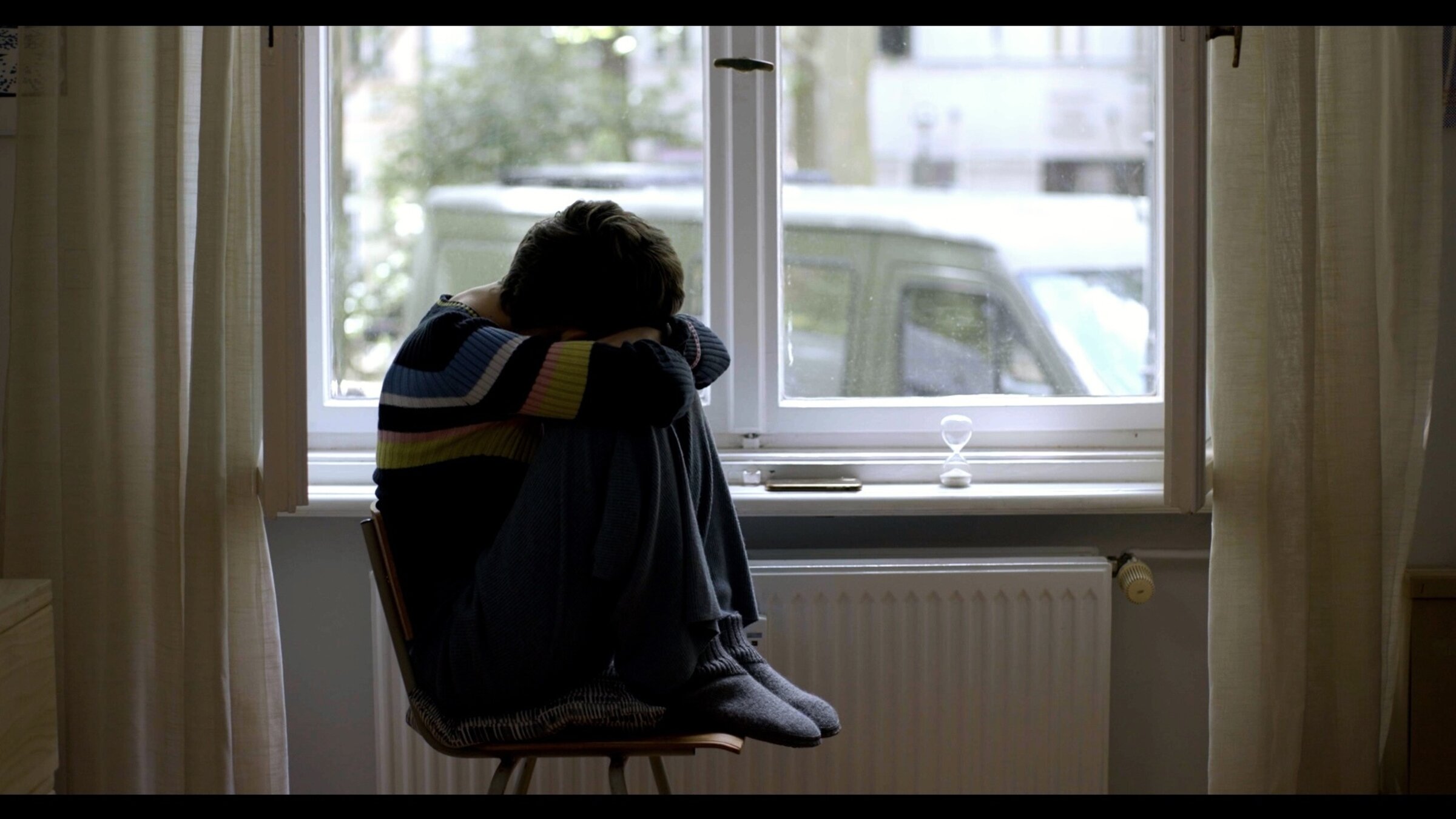 "DocuMe – Hello new body, how are you today?": Junge Frau sitzt mit angezogenen Beinen auf einem Stuhl an einem Fenster.