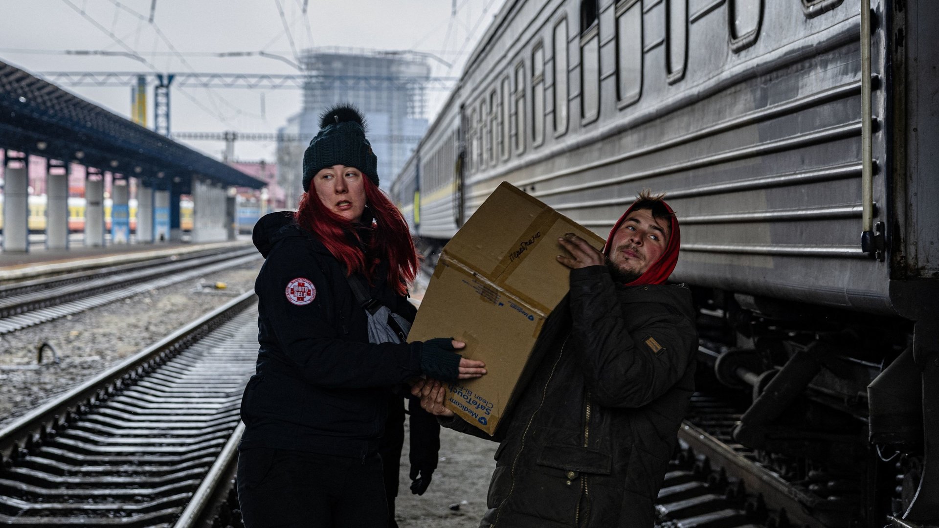 "Tatort Ukraine - Die Freiwilligen": Eine Frau und ein Mann tragen ein schweres Paket in einem menschenleeren Bahnhof über Bahngleis.