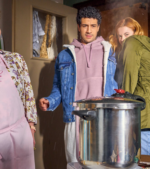 "Ich dich auch! - Der Unfall": Marion (Manon Straché), Yannik (Rojan Juan Barani) und Caro (Meriel Hinsching) stehen vor einem großen Schnellkochtopf, der auf dem Tisch steht. Skeptisch und besorgt dreinblickend weichen sie davor zurück.