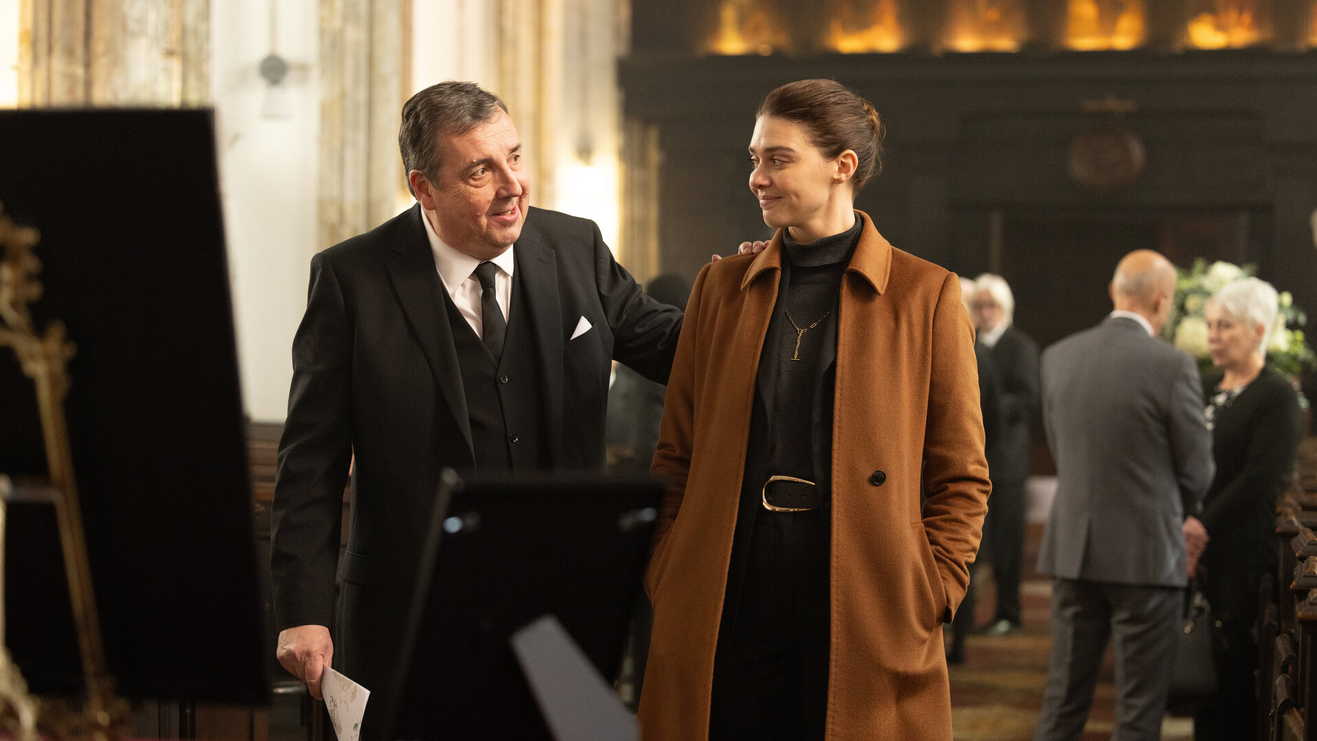 "The Killing Kind: Anonyme Grüße": Richter Stuart (Richard Dixon) und Ingrid Lewis (Emma Appleton) stehen während einer Trauerfeier an einem Sarg. Sie tragen formelle Kleidung und lächeln einander aufmunternd an, er hat außerdem seine linke Hand auf ihre Schulter gelegt. Im Hintergrund sind weitere Trauergäste (Komparsen) zu sehen.