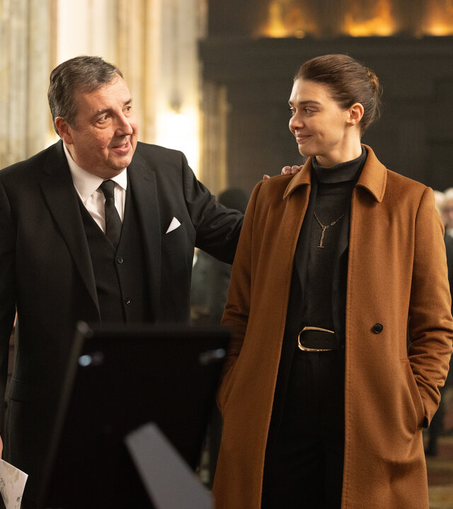 "The Killing Kind: Anonyme Grüße": Richter Stuart (Richard Dixon) und Ingrid Lewis (Emma Appleton) stehen während einer Trauerfeier an einem Sarg. Sie tragen formelle Kleidung und lächeln einander aufmunternd an, er hat außerdem seine linke Hand auf ihre Schulter gelegt. Im Hintergrund sind weitere Trauergäste (Komparsen) zu sehen.