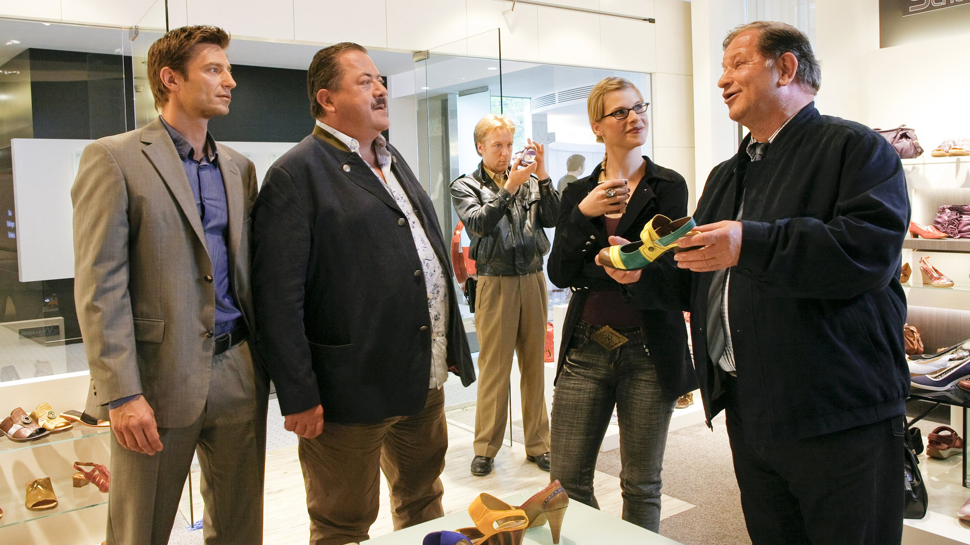 "Die Rosenheim-Cops: Der letzte Atemzug": Sven Hansen (Igor Jeftic), Korbinian Hofer (Joseph Hannesschläger), Michi Mohr (Max Müller), Lydia Seibold (Dagmar Geppert) und Schustermeister Josef Rabl (Anton Rattinger) befinden sich in einem Schuhgeschäft. Die beiden Kommissare schauen zu Rabl, der ihnen einen Damenschuh zeigt. Daneben steht lächelnd Lydia Seibold, die Rabl ansieht. Etwas abseits inspiziert Michi Mohr einen Gegenstand, den er nach oben hält.