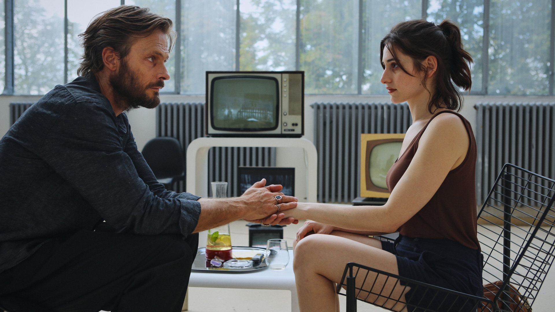"Der Schatten - Chaos": Wolfgang Balder (Andreas Pietschmann) sitzt auf einem Stuhl, vor ihm Norah Richter (Deleila Piasko). Balder hält ihre Hand. Im Hintergrund auf einem kleinen Tisch stehen eine Karaffe mit Wasser sowie alte Röhrenfernseher.