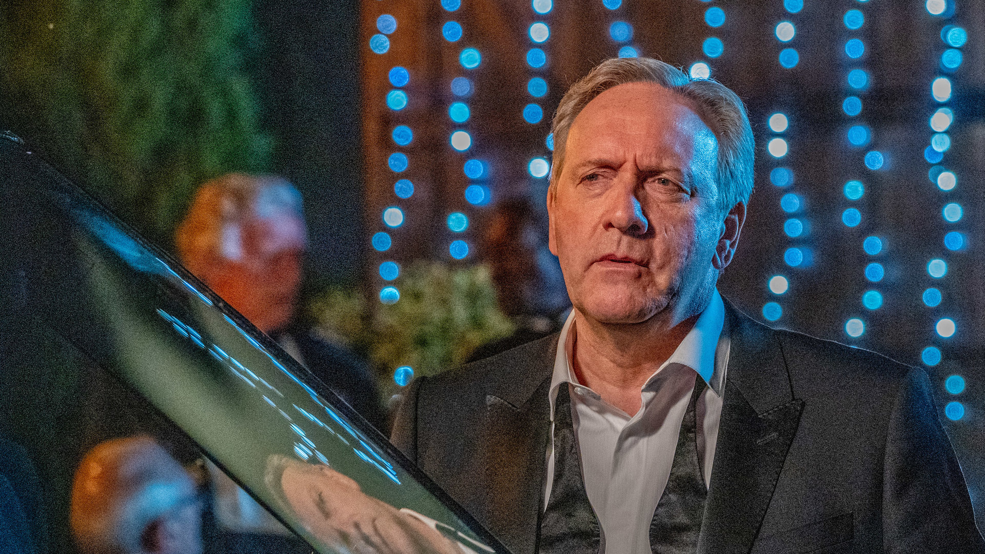 "Inspector Barnaby: Die Last der Lügen": DCI John Barnaby (Neil Dudgeon) in einem schwarzen Smoking gekleidet. Um seinen Hemdkragen hängt eine Fliege, steht vor einem Auto und schaut mit ernstem Blick. Im Hintergrund hängt eine blau leuchtende Lichterkette.