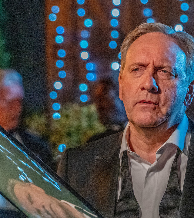 "Inspector Barnaby: Die Last der Lügen": DCI John Barnaby (Neil Dudgeon) in einem schwarzen Smoking gekleidet. Um seinen Hemdkragen hängt eine Fliege, steht vor einem Auto und schaut mit ernstem Blick. Im Hintergrund hängt eine blau leuchtende Lichterkette.