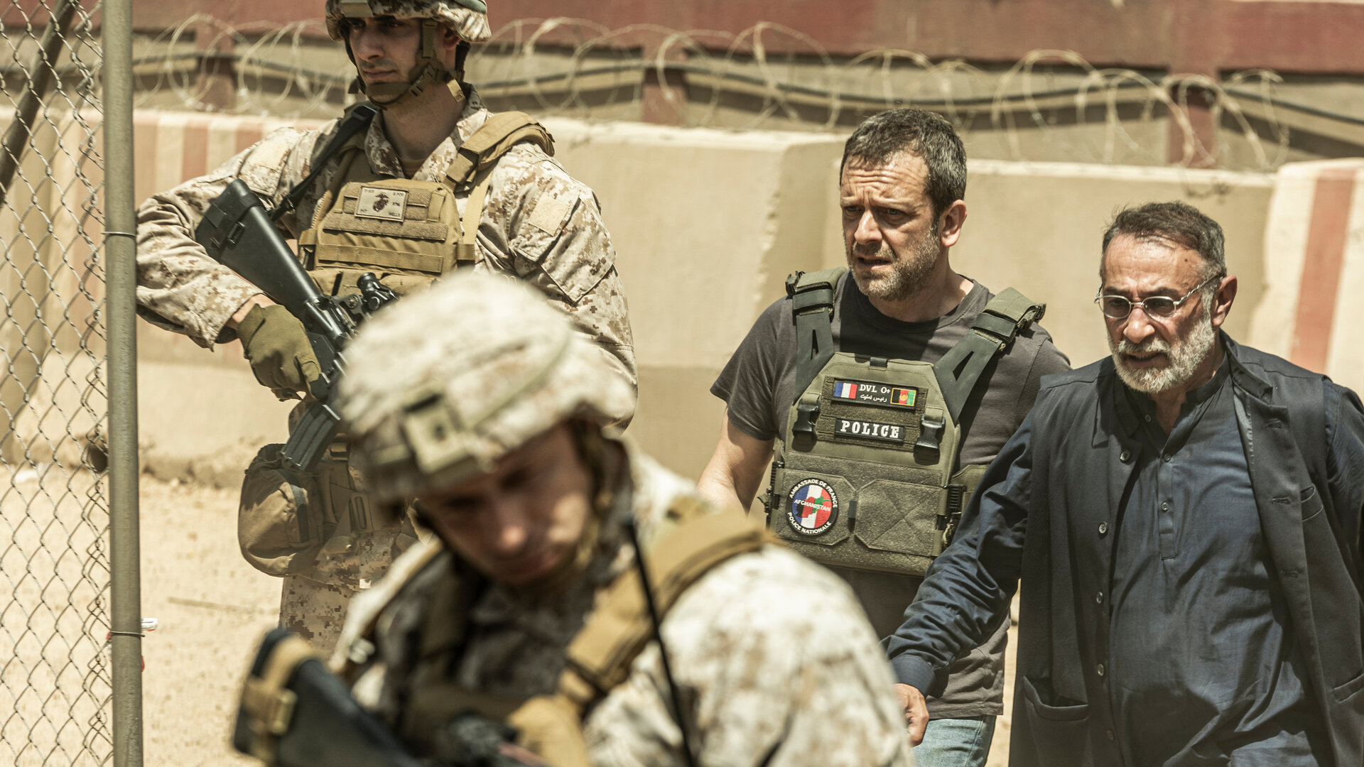 "Kabul - Flucht aus der Hölle: Alarmstufe Rot": Gilles (Jonathan Zaccaï) führt Baqir (Vassilis Koukalani) durch eine militärisch gesicherte Zone. Zwei amerikanische Soldaten in voller Kampfmontur (unbekannte Darsteller) überwachen die Umgebung, die Situation wirkt angespannt und gefährlich.