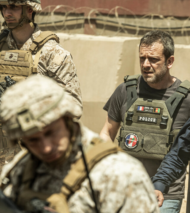 "Kabul - Flucht aus der Hölle: Alarmstufe Rot": Gilles (Jonathan Zaccaï) führt Baqir (Vassilis Koukalani) durch eine militärisch gesicherte Zone. Zwei amerikanische Soldaten in voller Kampfmontur (unbekannte Darsteller) überwachen die Umgebung, die Situation wirkt angespannt und gefährlich.