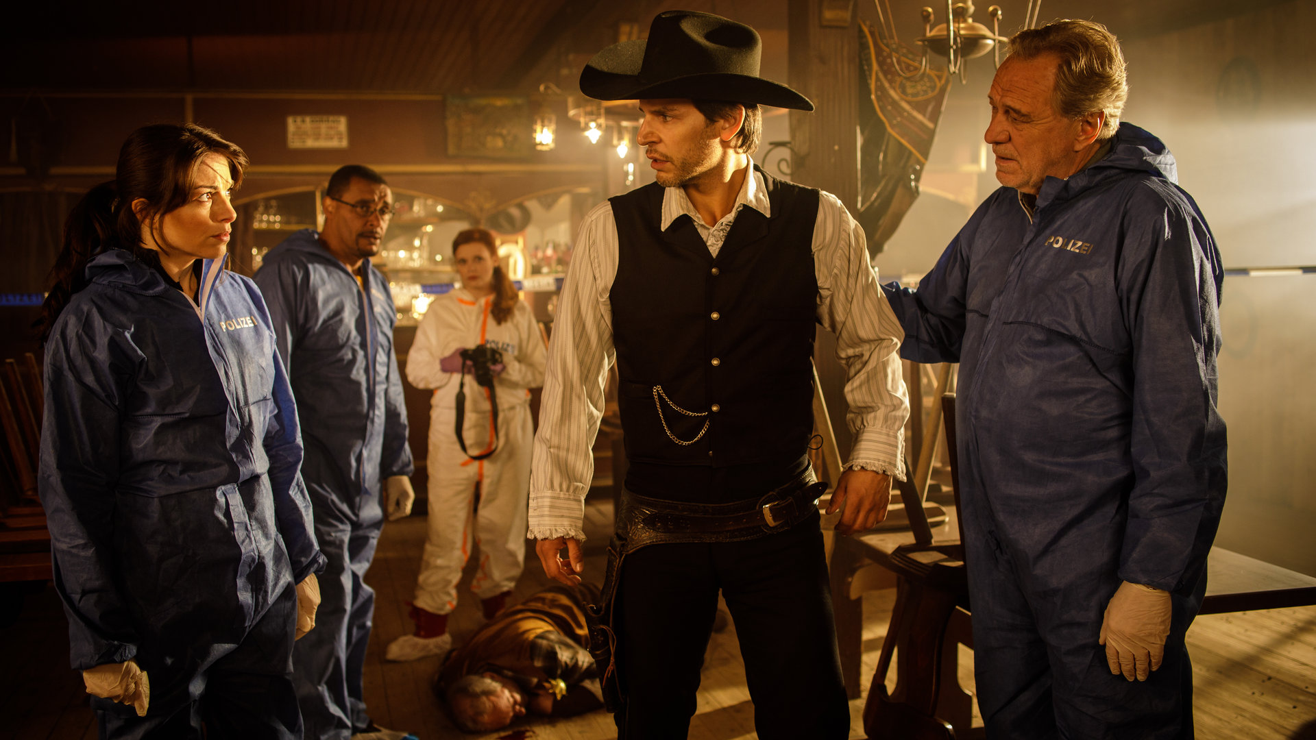 "SOKO München - Himmel Herrgott, Sacramento": In einem Saloon stehen Katharina Hahn (Bianca Hein), Franz Ainfachnur (Christofer v. Beau) und Billie Curio (Sina Reiß). Sie schauen zu Dr. Weissenböck (Florian Odendahl), der als Cowboy verkleidet ist. Hinter ihm steht Arthur Bauer (Gerd Silberbauer) und legt ihm die Hand auf den Rücken. Auf dem Boden liegt eine Leiche.