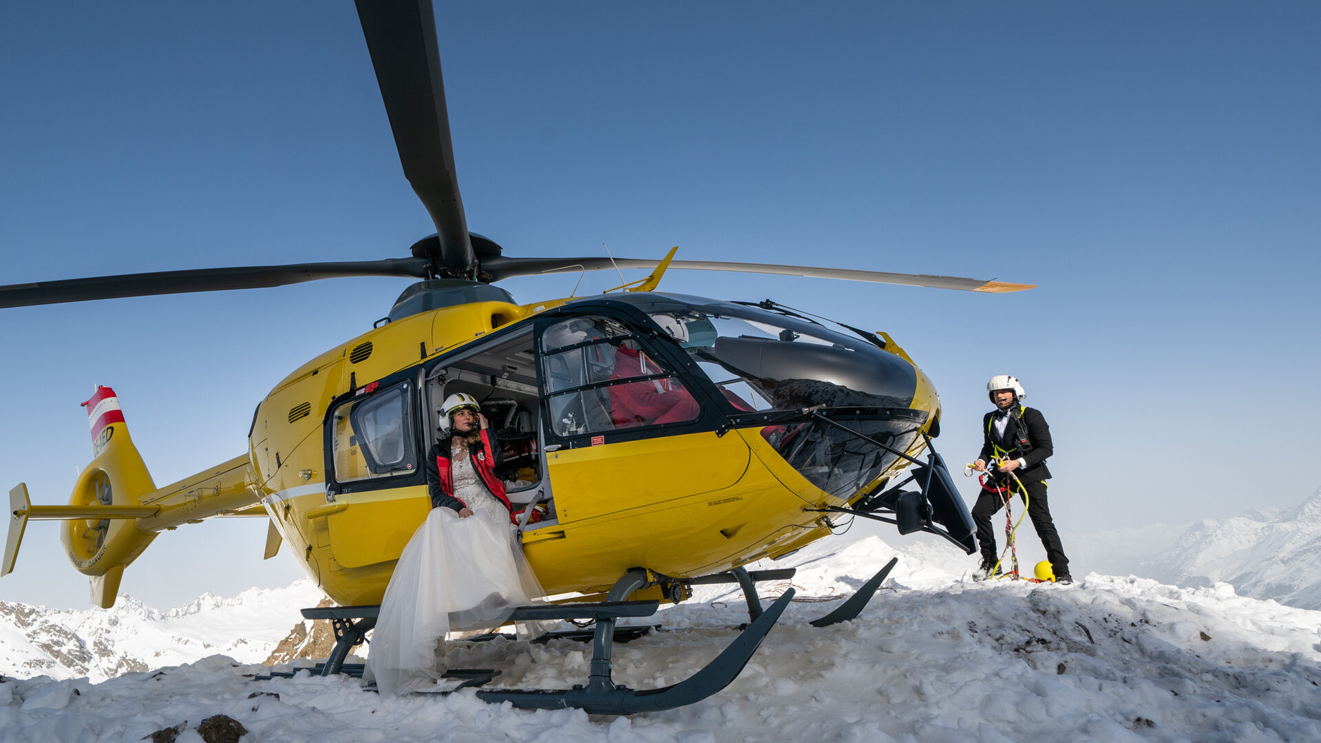 "Die Bergretter: Dieses eine Leben": Der Rettungshubschrauber steht auf einer schneebedeckten Bergspitze. In der geöffneten Tür sitzt Katharina (Luise Bähr), die ein weißes Brautkleid trägt. Markus (Sebastian Ströbel) steht in einem Anzug etwas abseits.