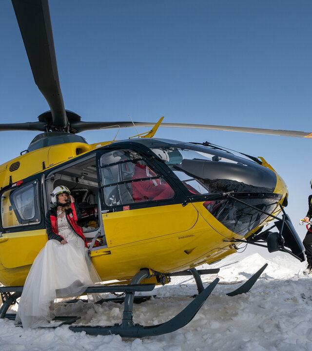 "Die Bergretter: Dieses eine Leben": Der Rettungshubschrauber steht auf einer schneebedeckten Bergspitze. In der geöffneten Tür sitzt Katharina (Luise Bähr), die ein weißes Brautkleid trägt. Markus (Sebastian Ströbel) steht in einem Anzug etwas abseits.