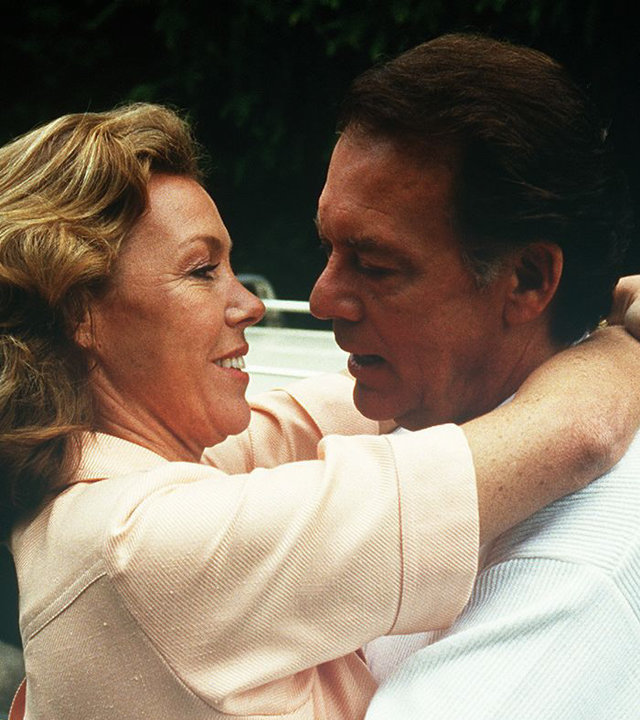 "Die Schwarzwaldklinik - Die Erbschaft": Christa (Gaby Dohm) und Klaus Brinkmann (Klausjürgen Wussow) liegen sich glücklich in den Armen und schauen sich in die Augen. Im Hintergrund ist ein Auto zu sehen.