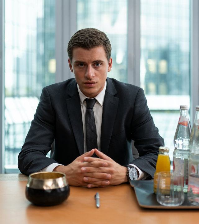 "Füxe - Den Tüchtigen hilft das Glück": Adem (Valon Krasniqi) sitzt, in schwarzem Anzug und Krawatte gekleidet, an einem Besprechungstisch. Vor ihm stehen Wasserflaschen und kleine Saftflaschen.