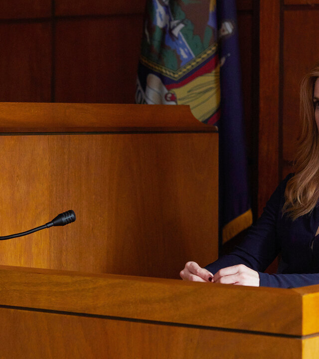 "Suits - Donna": Donna (Sarah Rafferty) sitzt im Zeugenstand in einem Gerichtssaal. Sie schaut konzentriert. Ihre Hände sind auf dem Tisch abgelegt und berühren sich. Im Hintergrund ist eine Flagge zu erkennen.