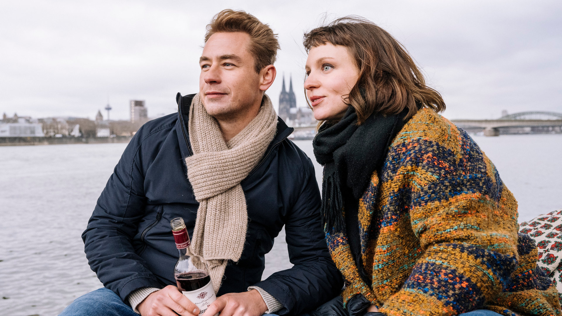 "SOKO Köln - Hallo Fremder": Maren Hofstätter (Maditha Dolle) und Kai Schimmel (Niklas Löffler) sitzen nebeneinander am Rheinufer und blicken lächelnd in die Ferne, links an der Kamera vorbei. Sie lehnen sich dabei zueinander. Niklas hält eine Flasche mit einem dunkelroten Inhalt in der Hand. Der Himmel ist bedeckt.