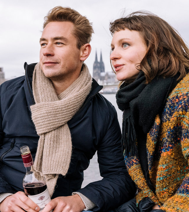"SOKO Köln - Hallo Fremder": Maren Hofstätter (Maditha Dolle) und Kai Schimmel (Niklas Löffler) sitzen nebeneinander am Rheinufer und blicken lächelnd in die Ferne, links an der Kamera vorbei. Sie lehnen sich dabei zueinander. Niklas hält eine Flasche mit einem dunkelroten Inhalt in der Hand. Der Himmel ist bedeckt.