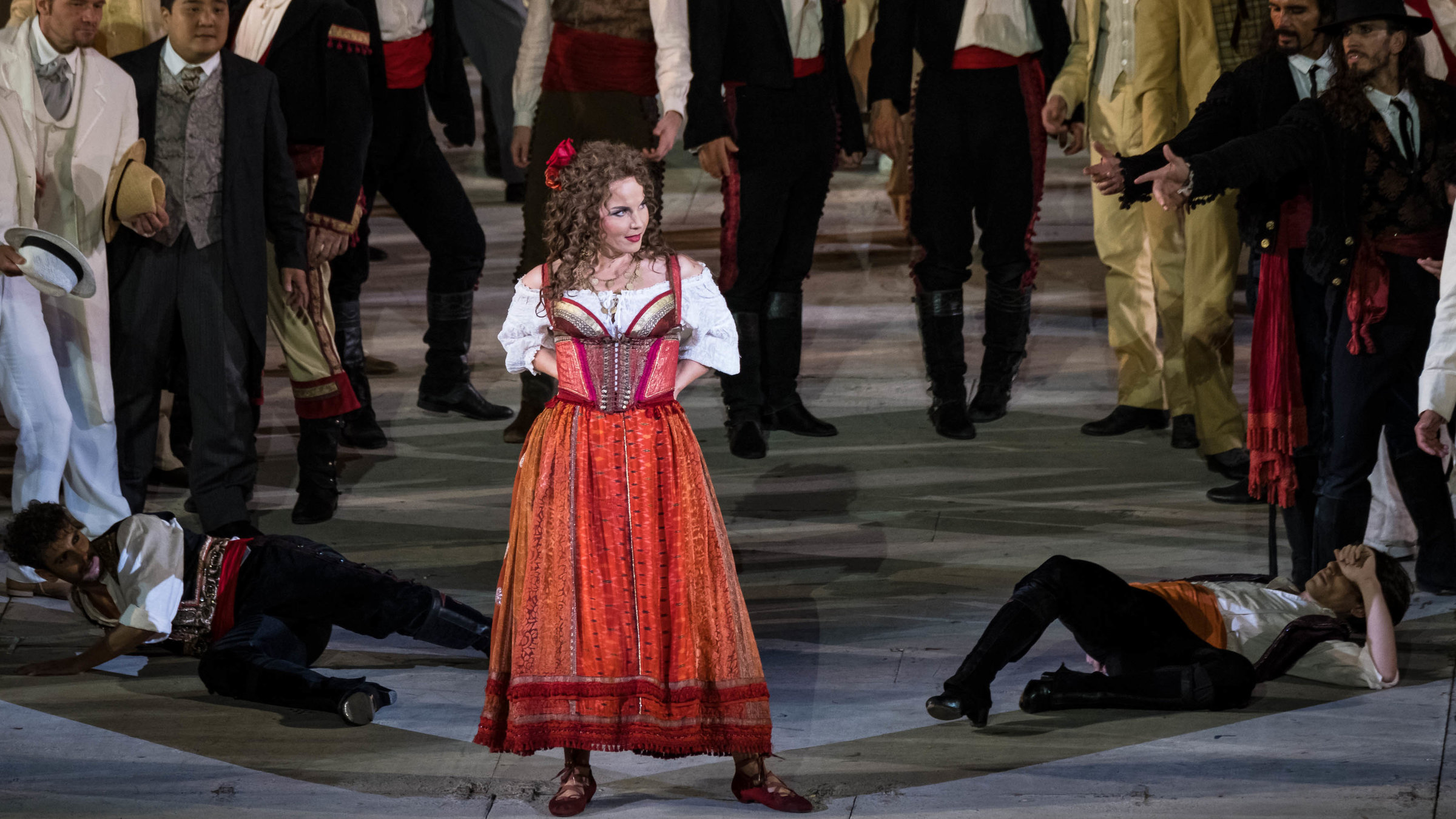 "Aus der Arena di Verona - Georges Bizet: Carmen": Carmen steht auf einer Bühne, umringt von vielen Männern. Sie hat die Hände in die Hüften gestemmt und blickt lächelnd zur Seite. Gekleidet ist sie mit einem trachtenartigen roten Kleid und einer weißen Bluse.