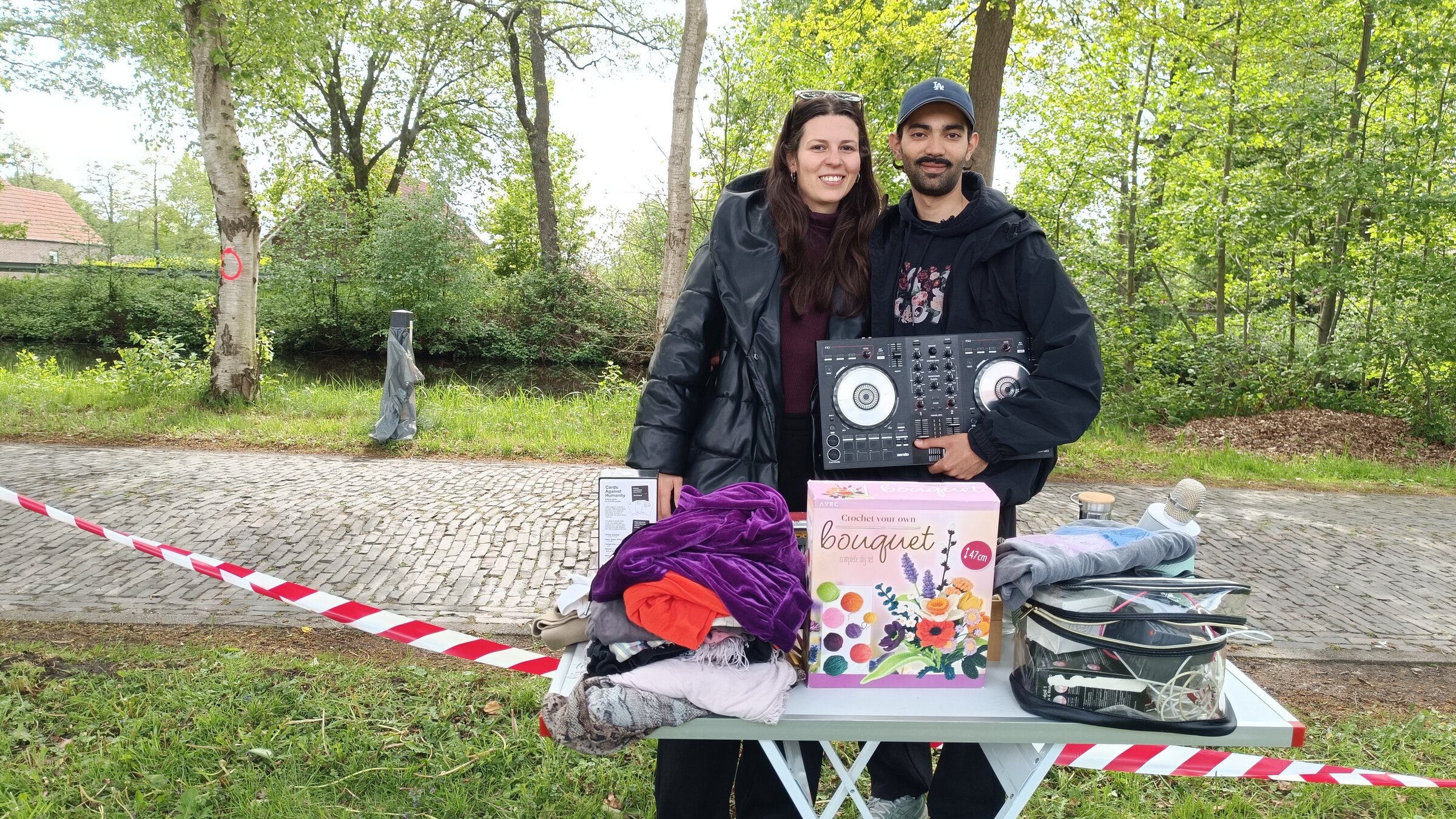 "ZDF.reportage: Flohmarkt XXL - Deutschlands längster Flohmarkt": Sabina und Ayhan stehen an einem Weg im Grünen. Vor ihnen steht ihr kleiner Flohmarktstand, ein Tisch auf dem verschiedene Sachen liegen.