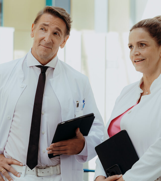 "Der Bergdoktor: Wünsche": In der Klinik: Dr. Vera Fendrich (Rebecca Immanuel) und Dr. Alexander Kahnweiler (Mark Keller) stehen nebeneinander im Flur und blicken überrascht nach vorne. Beide tragen ihren Arztkittel und halten Tablets in den Händen. Im Hintergrund läuft Pflegepersonal (Komparsen) durch den Flur.