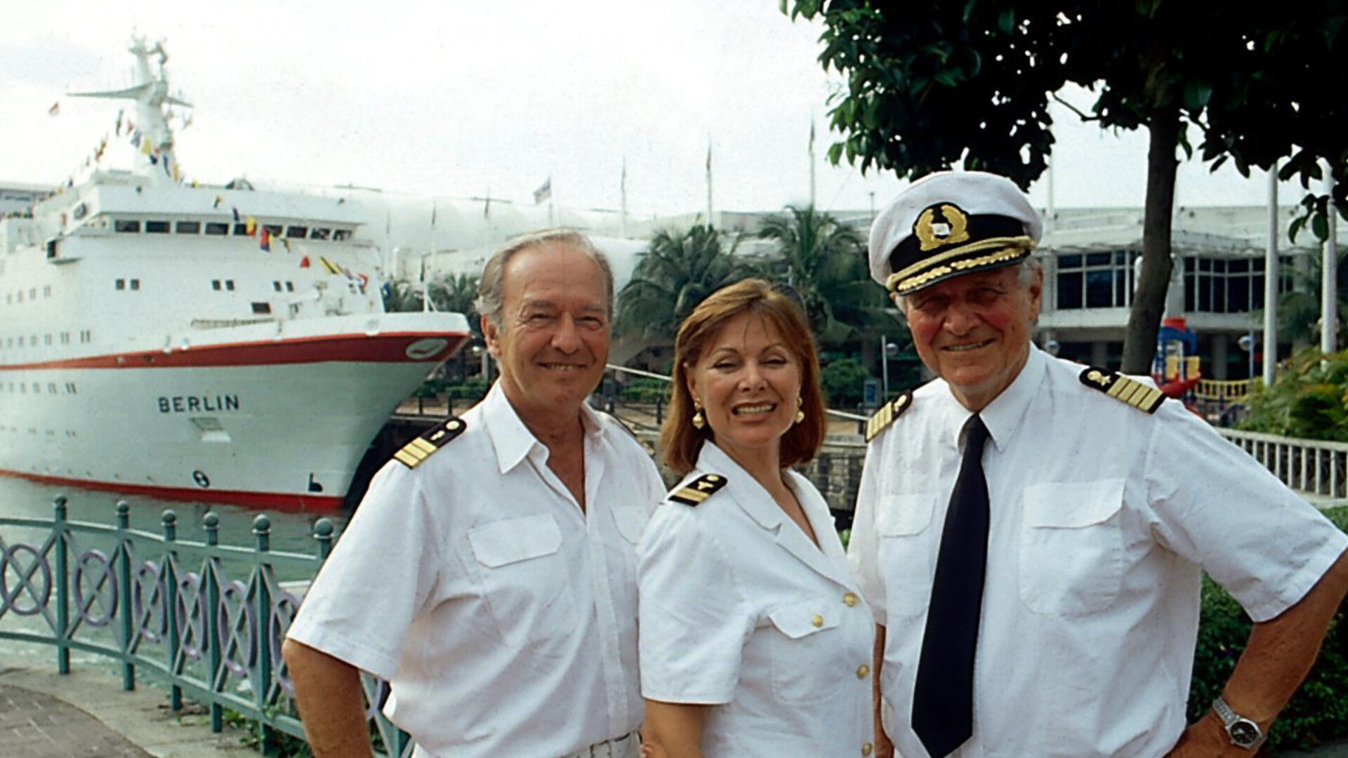 "Das Traumschiff: Singapur": Kapitän Hansen (Heinz Weiss), Chefhostess Beatrice (Heide Keller) und Schiffsarzt Dr. Horst Schröder (Horst Naumann) stehen in Uniform nebeneinander vor dem Traumschiff und lächeln fröhlich.