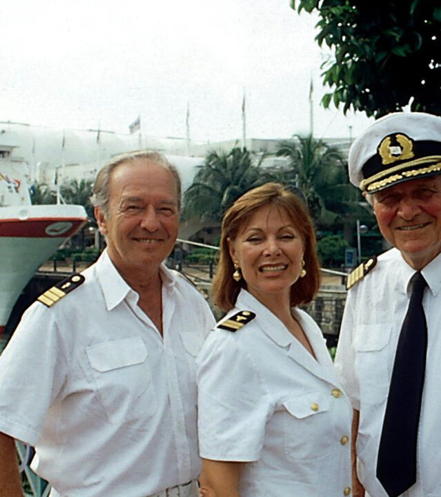 "Das Traumschiff: Singapur": Kapitän Hansen (Heinz Weiss), Chefhostess Beatrice (Heide Keller) und Schiffsarzt Dr. Horst Schröder (Horst Naumann) stehen in Uniform nebeneinander vor dem Traumschiff und lächeln fröhlich.