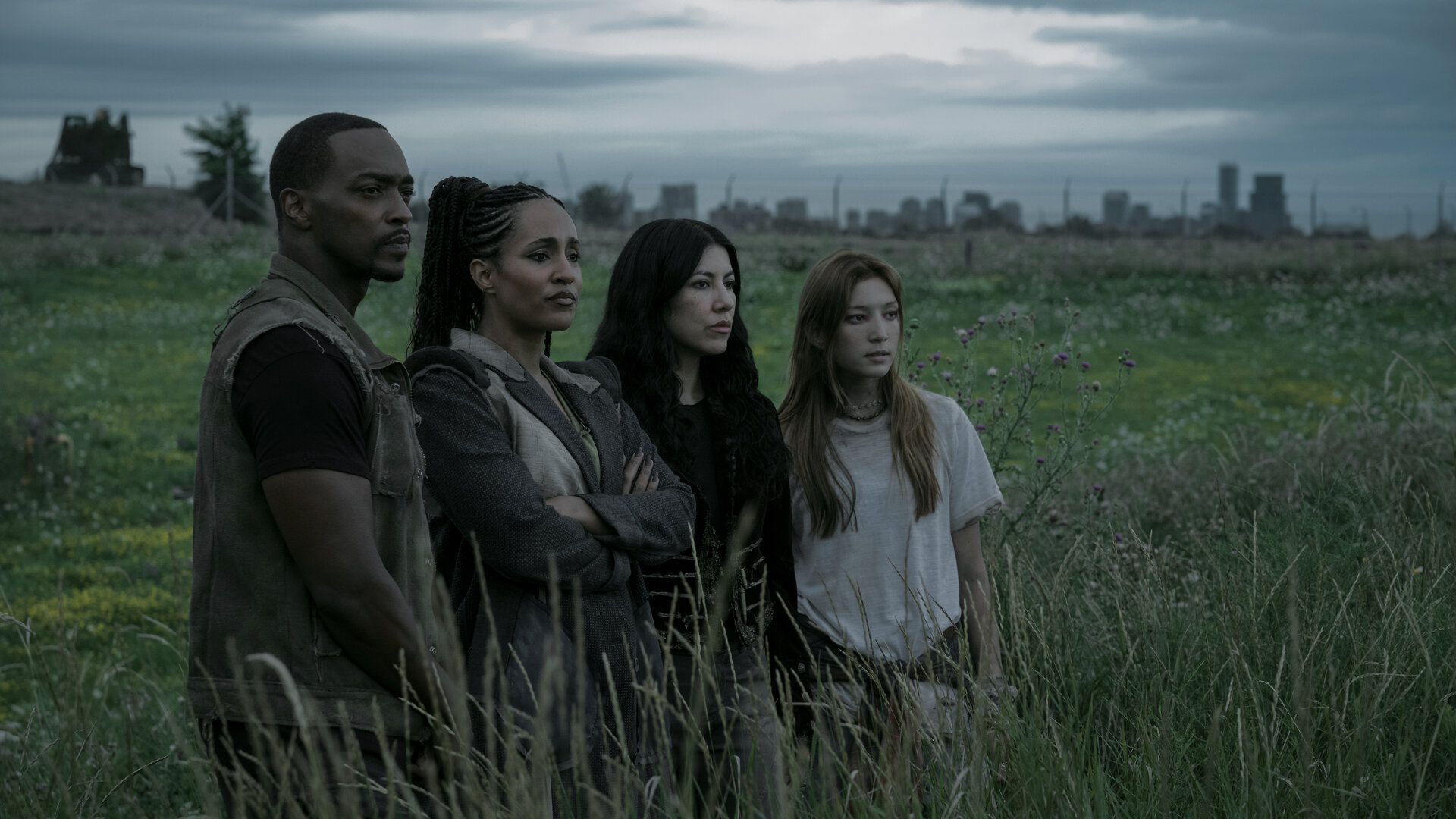 "Twisted Metal - T3STDRV": John (Anthony Mackie), Dollface (Tiana Okoye), Quiet (Stephanie Beatriz) und Mayhem (Saylor Bell Curda) stehen auf einer weiten grünen Wiese mit hohem Gras. Alle blicken mit ernstem Gesichtsausdruck zur Seite in dieselbe Richtung.